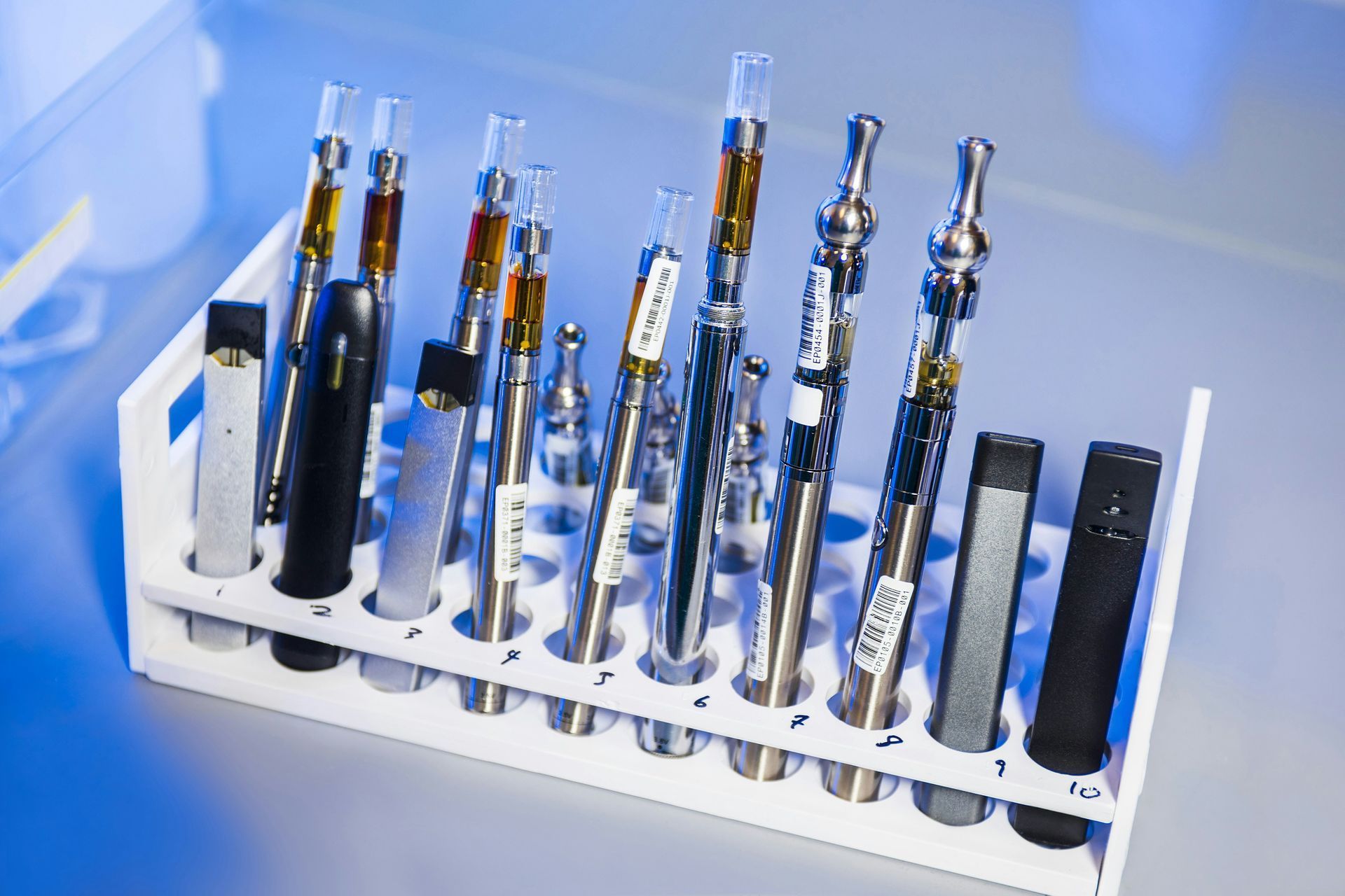 Vaping devices in a white rack. Some are black, silver, and clear, filled with brown liquid.