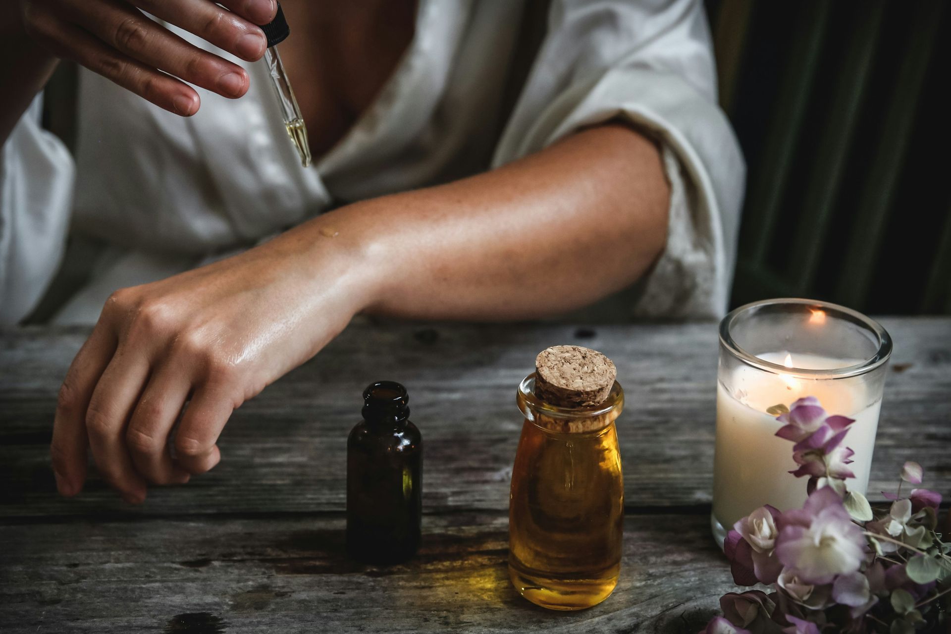 Person applying oil to arm from dropper, bottles and candle on a wooden table.
