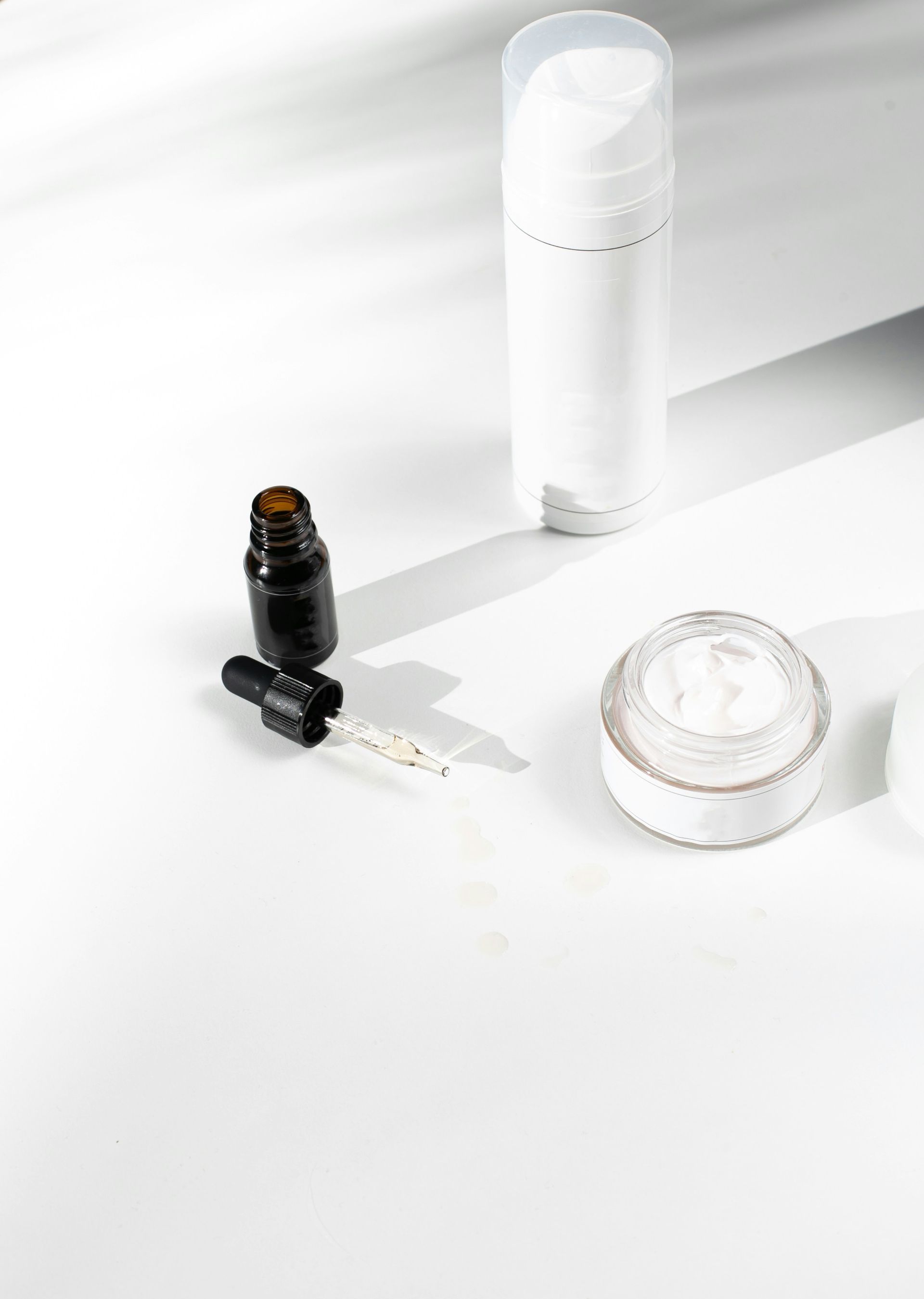 Skincare products on a white surface: jar of cream, pump bottle, dropper bottle with dropper.