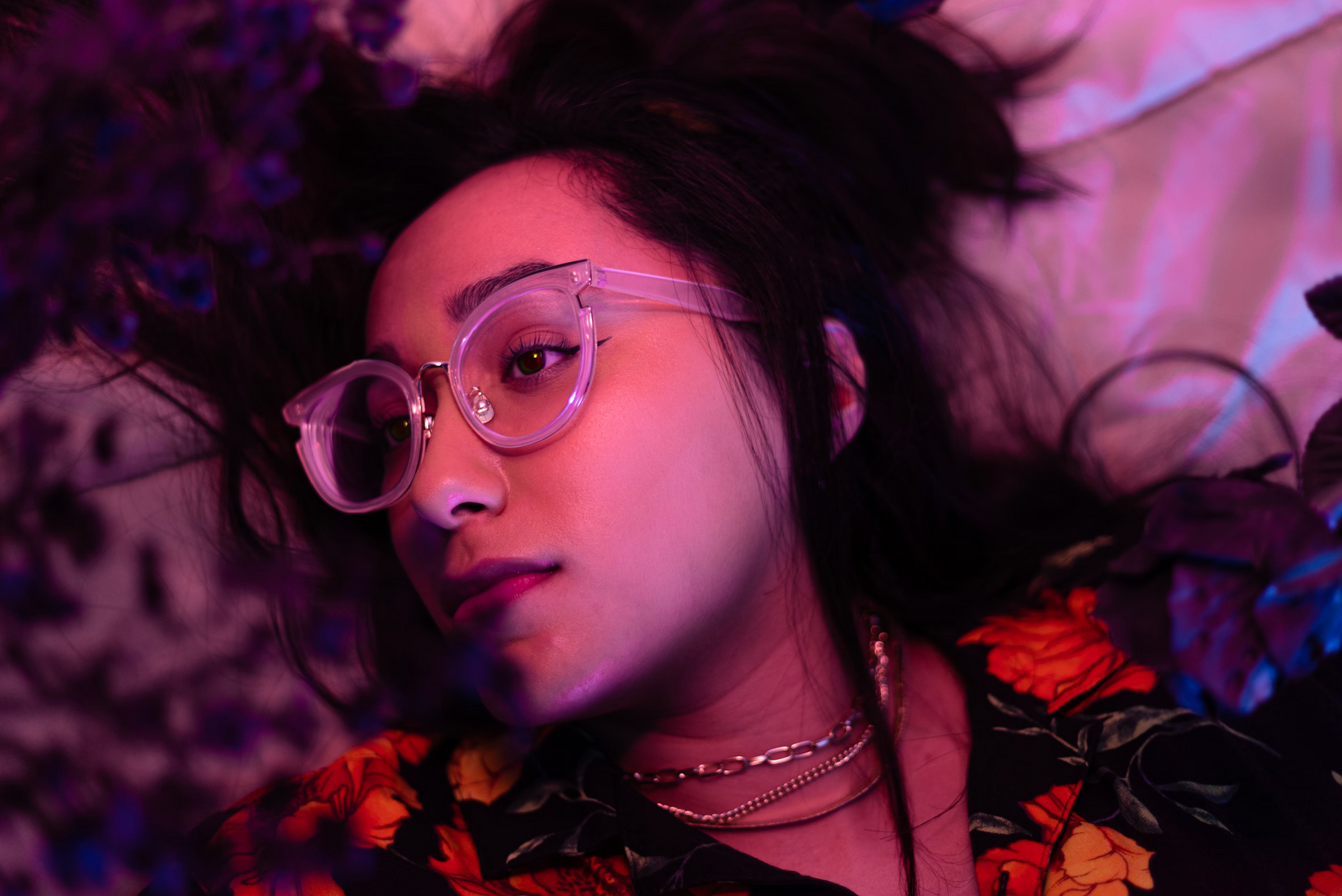 Woman wearing glasses, lying down, looking to the side with flowers in a dimly lit setting.