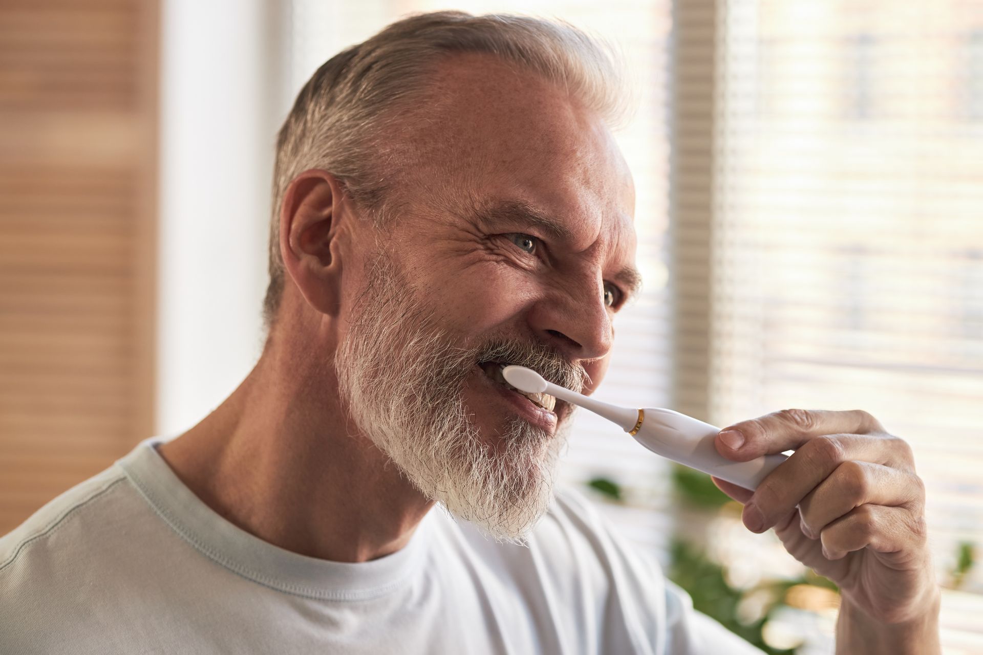Man brushing his teeth with a white toothbrush in a bright bathroom, smiling slightly.