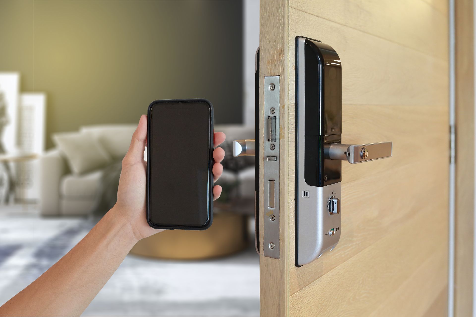 Hand holding a smartphone next to a modern smart door lock installed on a wooden door.