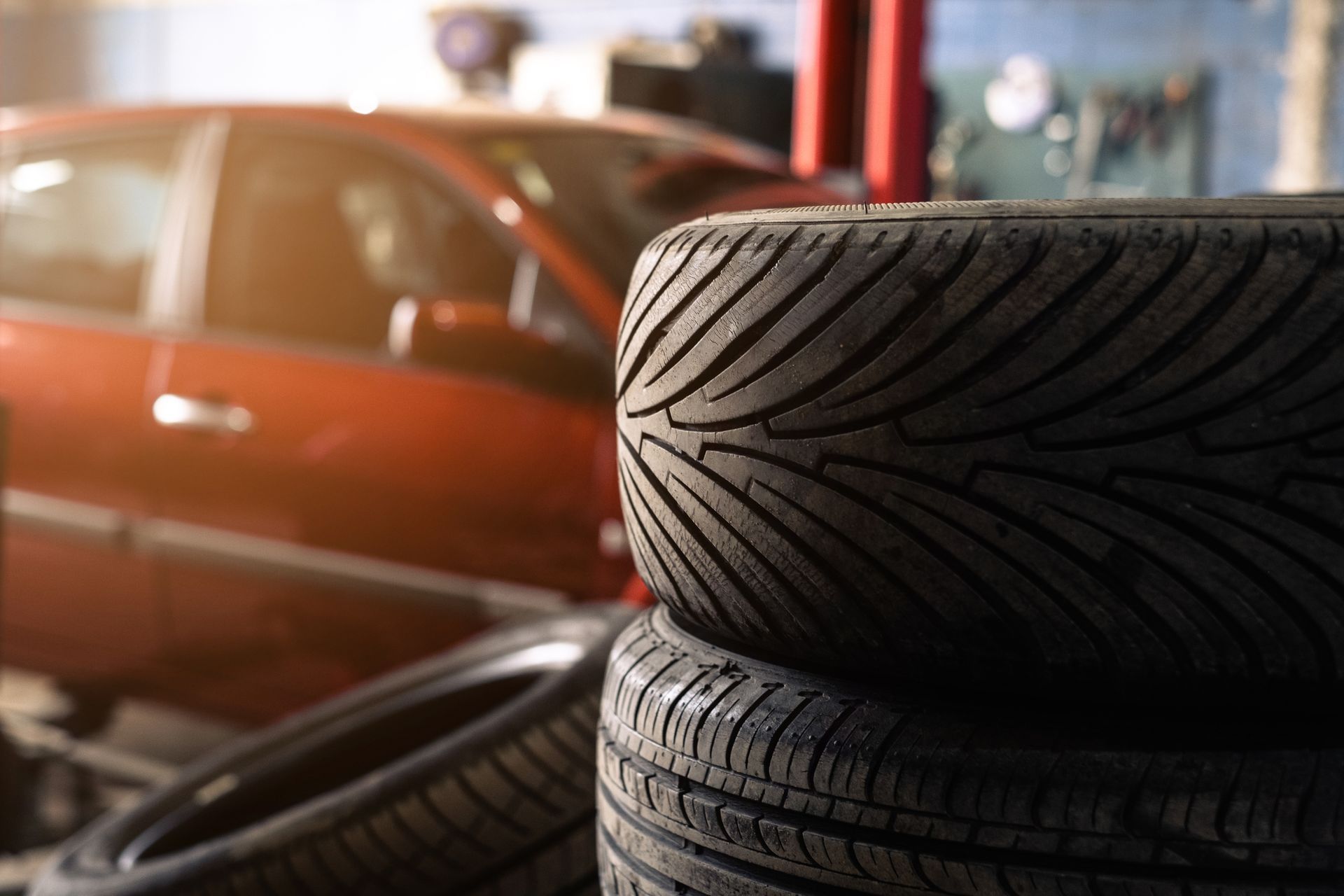 Stack of car tires in a garage with a red car in the background.