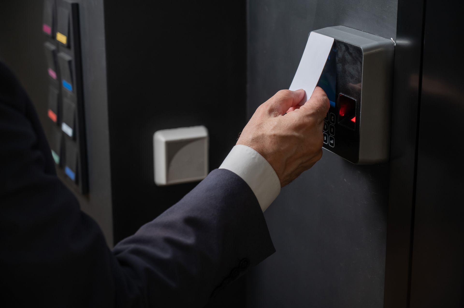 A person in a dark suit holds a white card against an electronic access control reader on a wall.