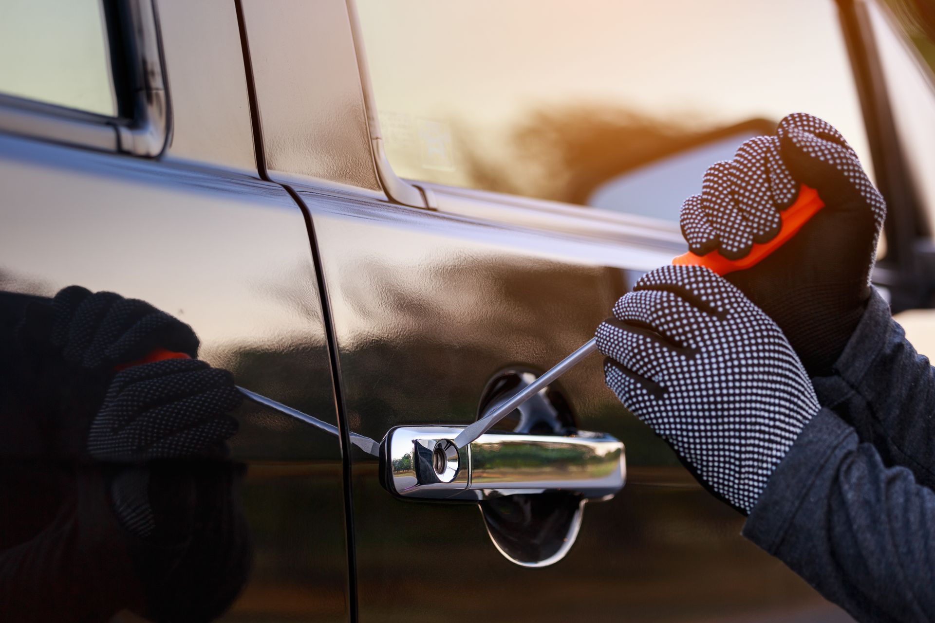 A person wearing patterned gloves uses a screwdriver to pry open the door handle of a dark-colored vehicle.