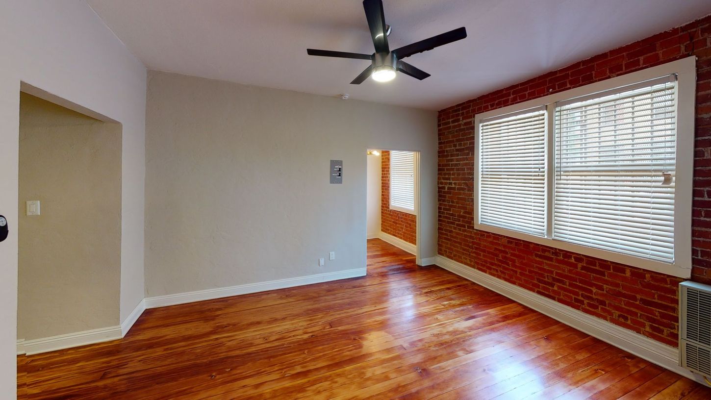 Empty room with wood floors, exposed brick wall, window, ceiling fan, and open doorway.