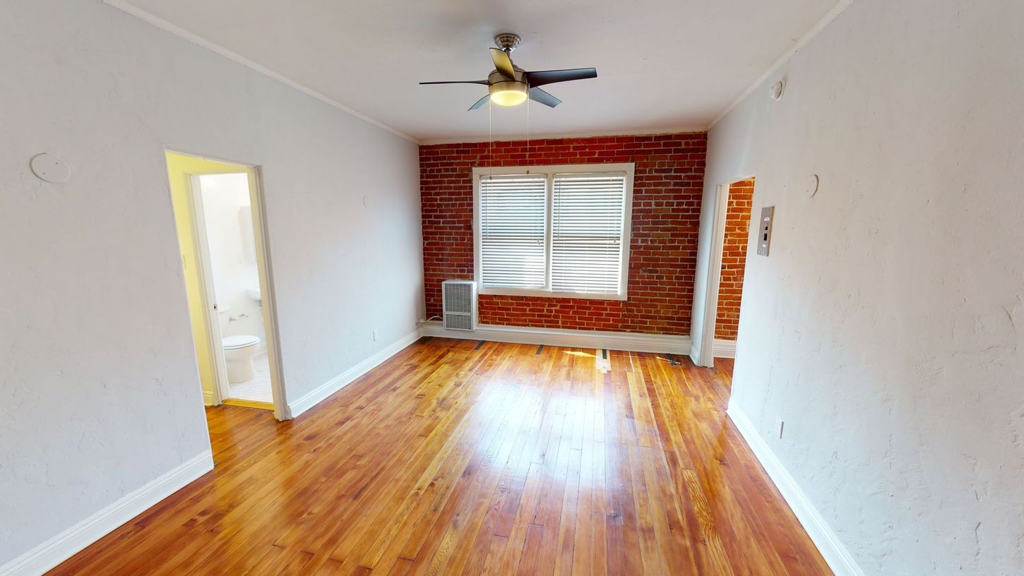 Interior room with brick wall, wood floors, and ceiling fan. White walls, window with blinds.