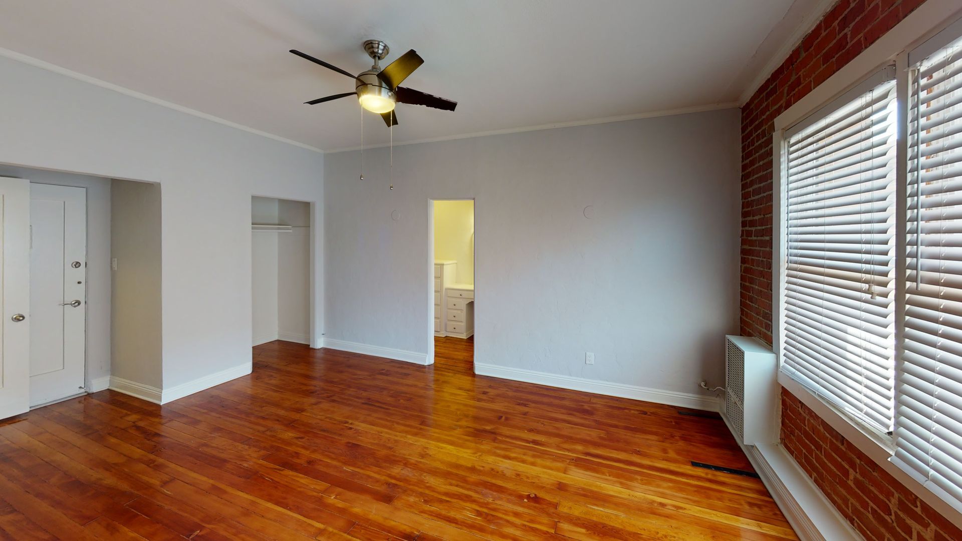 Interior of a room with wood floors, exposed brick, a window with blinds, and a ceiling fan.