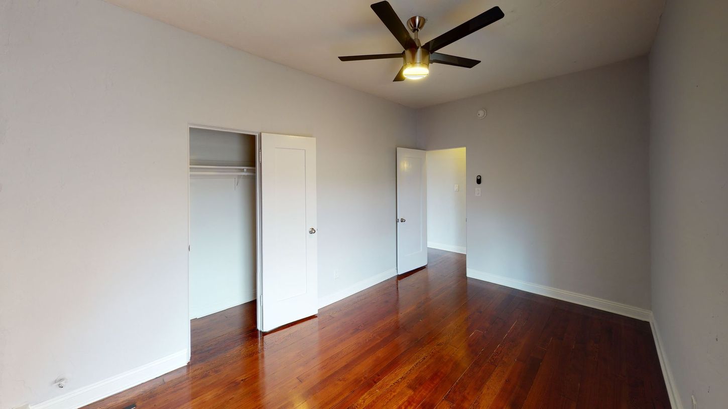 Empty bedroom with hardwood floors, two white doors, closet, and ceiling fan. Gray walls.