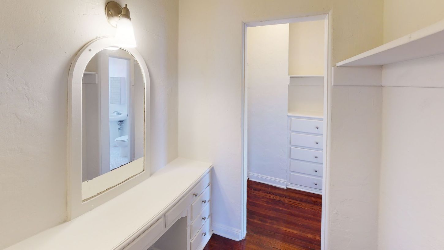 White vanity with mirror, overhead light, closet entrance, and wooden floor.