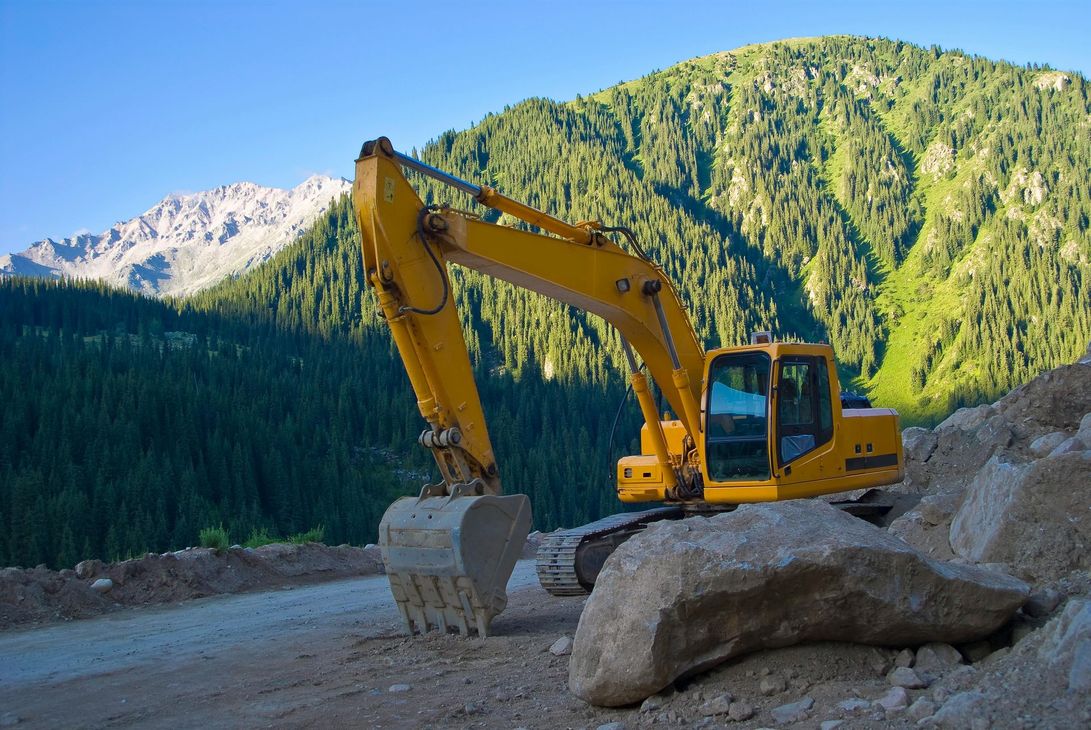 Yellow excavator on a dirt road next to rocks, mountain backdrop.