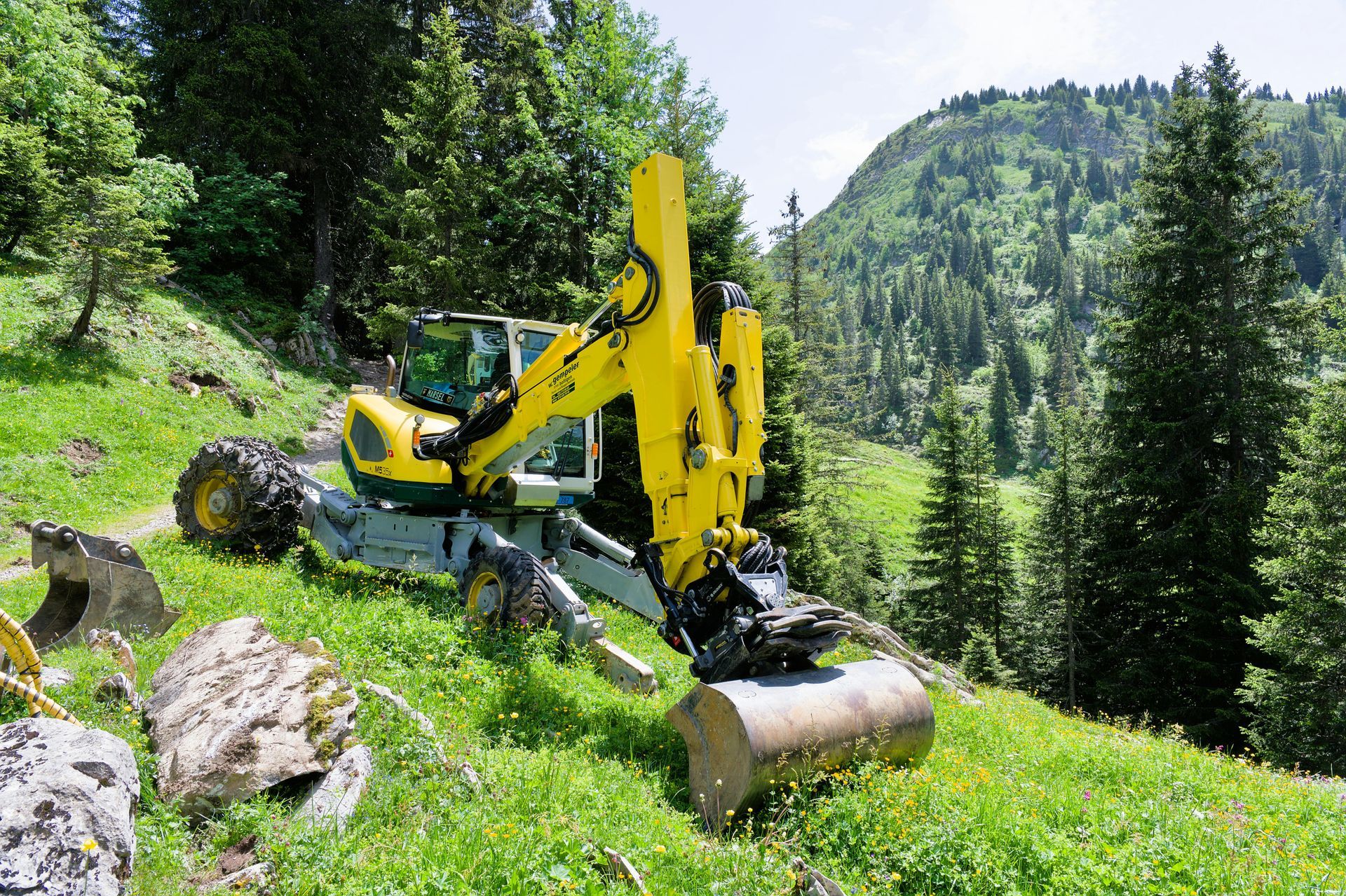 Yellow excavator working on a grassy hillside, surrounded by trees, mountains in the background.