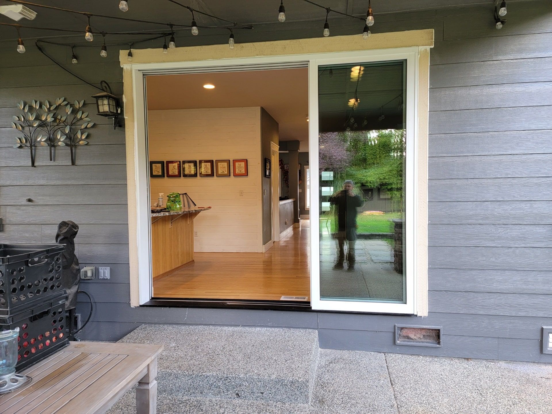 Open sliding glass door to interior, reflecting a person. Outdoor patio, gray siding, string lights.