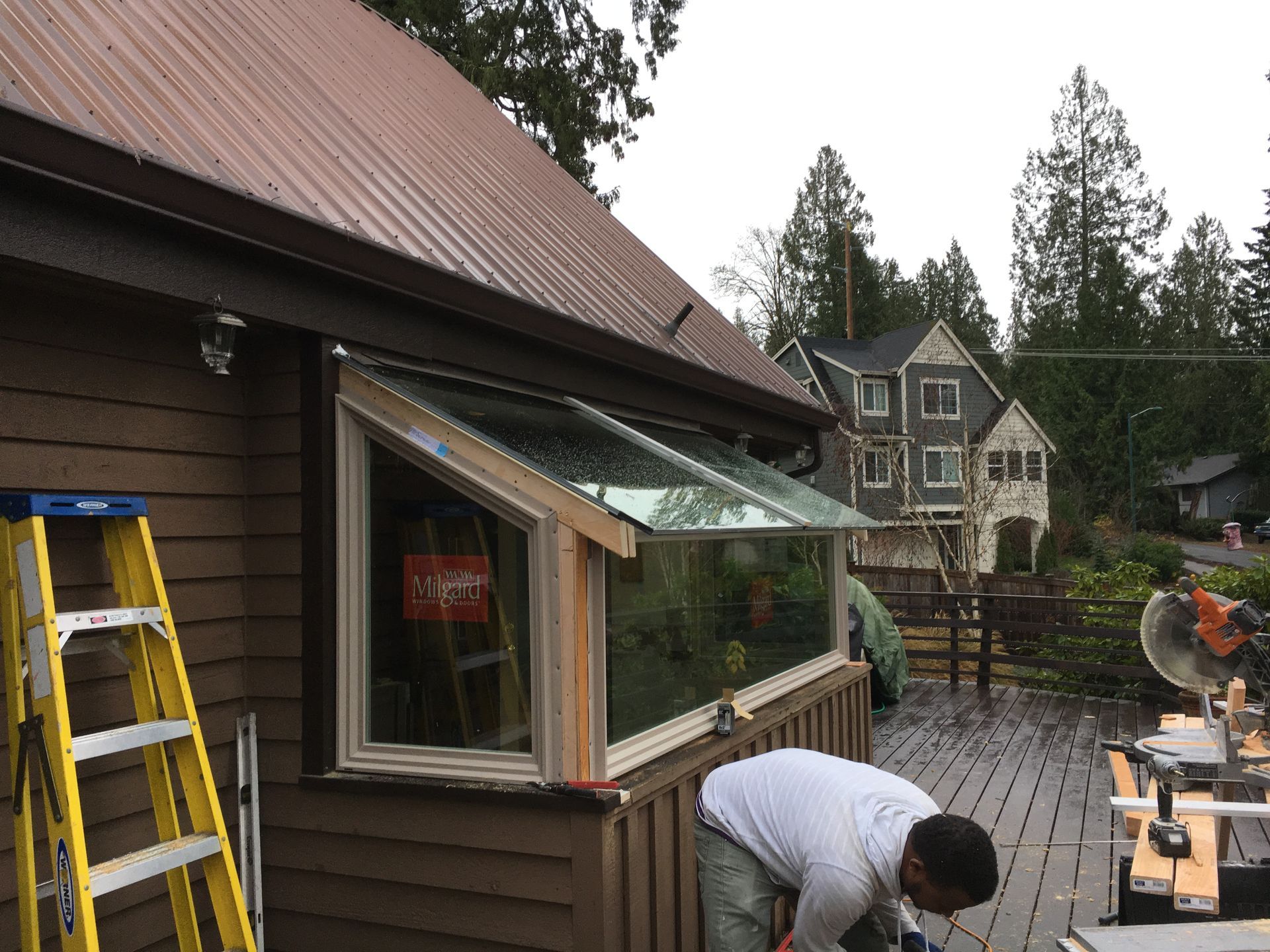 Man working on a deck near a building with a window and metal roof. A yellow ladder stands nearby.