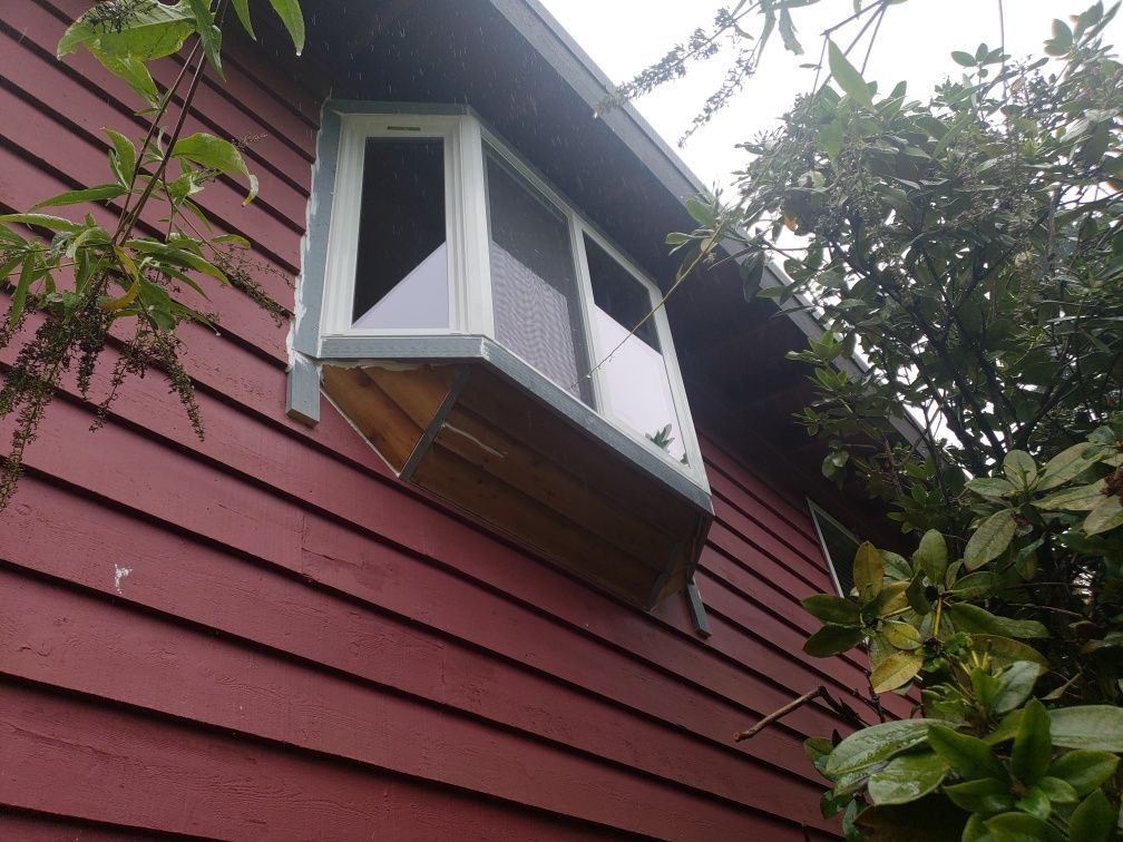 Bay window on a red house, with a wooden box beneath. Green foliage surrounds it.