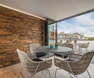 Balcony with wood-paneled wall, round table and woven chairs, open to an outdoor dining area with city view.