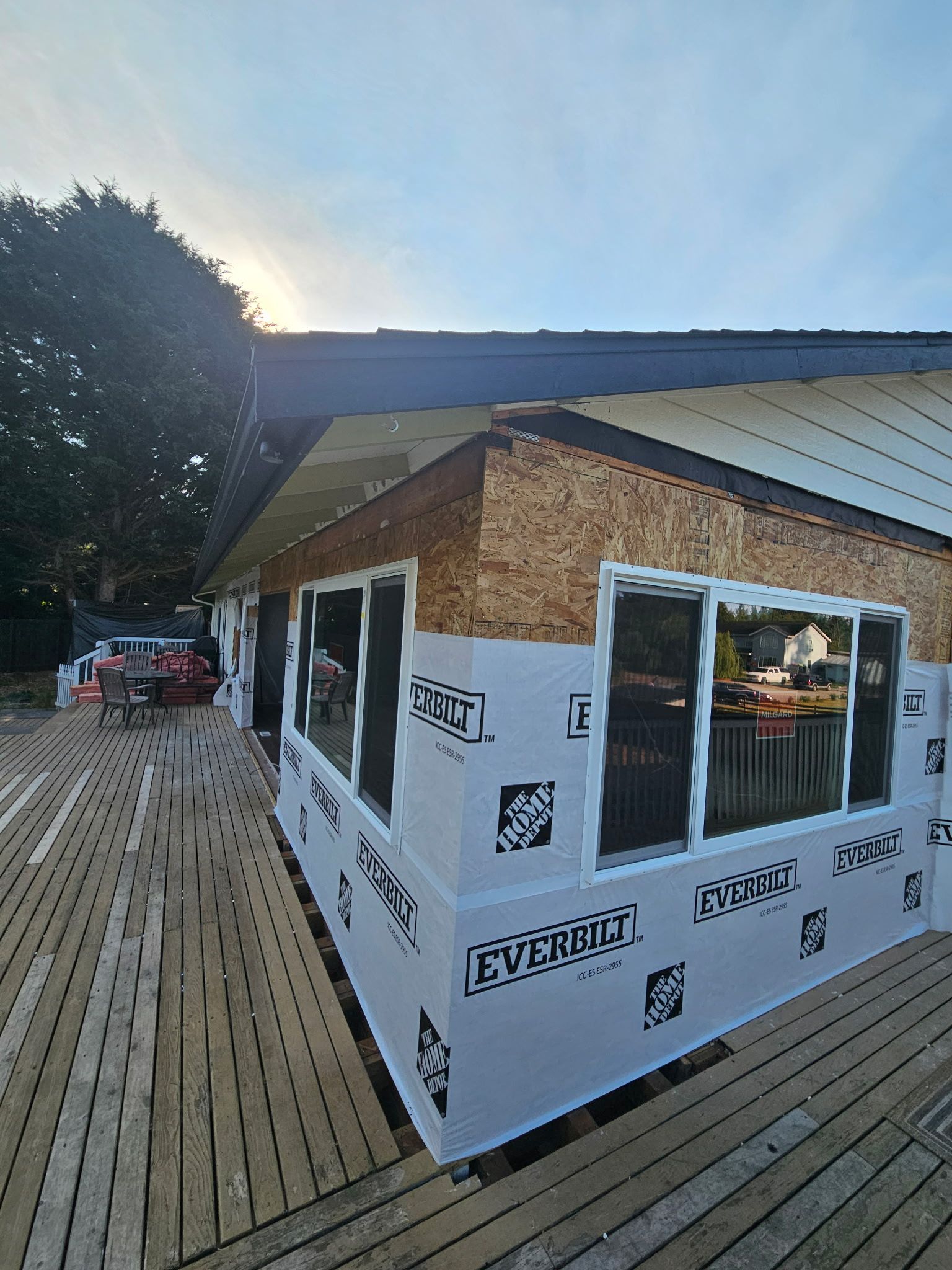 House under construction, with exposed wood siding and windows; wrapped in white material, deck in foreground.
