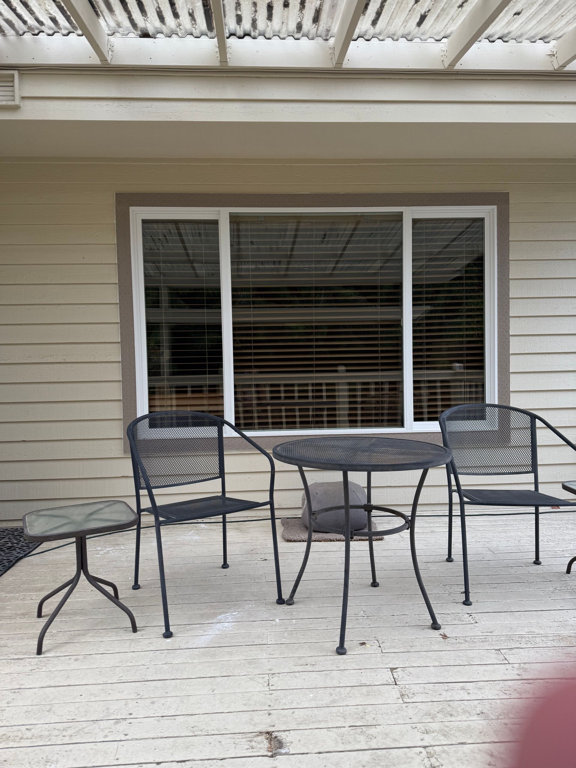 Outdoor patio with metal furniture in front of a window with blinds. Beige siding and pergola overhead.
