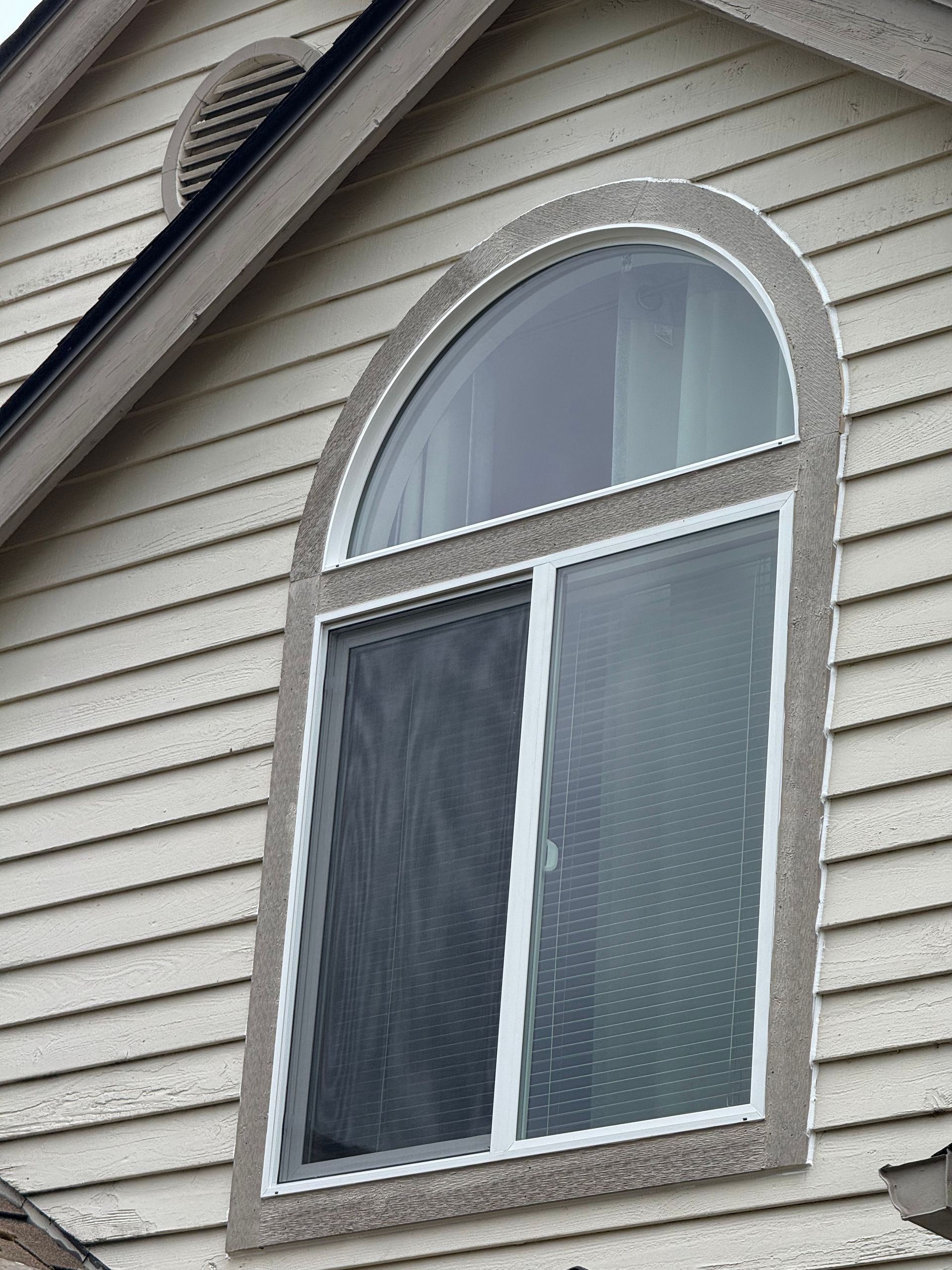 Arched window with a screen, set in tan siding. Above, a small circular vent on a beige wall.