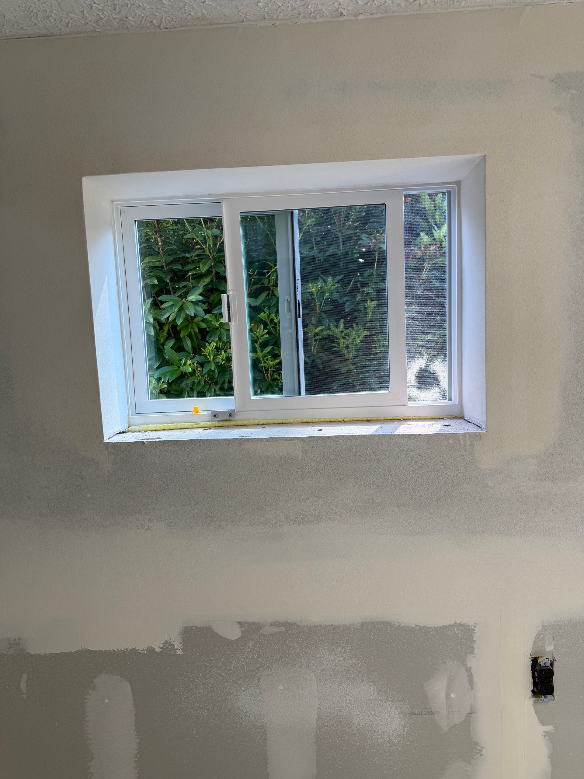 Window with a white frame in a room with unfinished walls, view of green foliage outside.