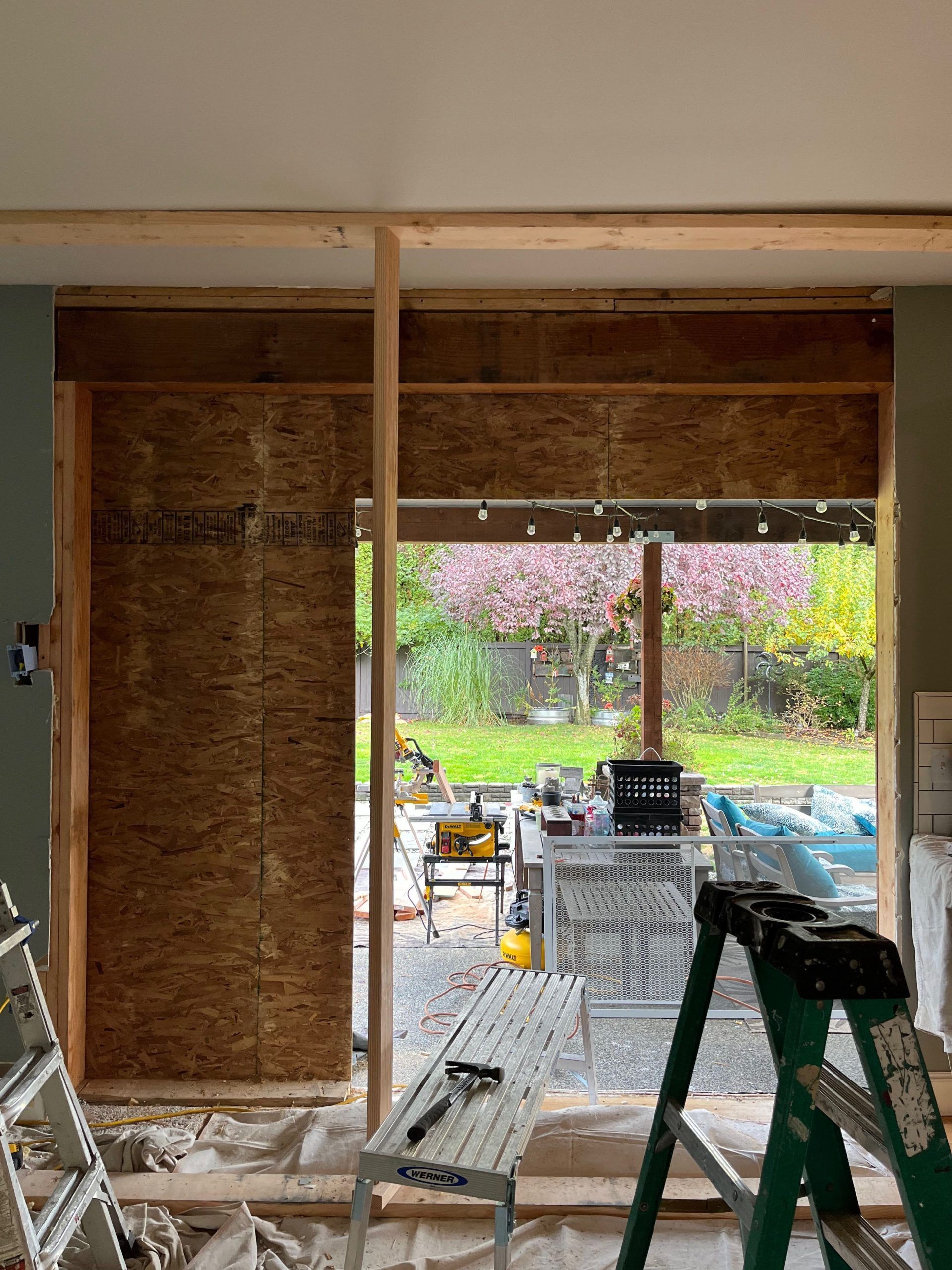 Construction site: doorway frame with exposed insulation, looking out to a garden. Tools and ladder are present.