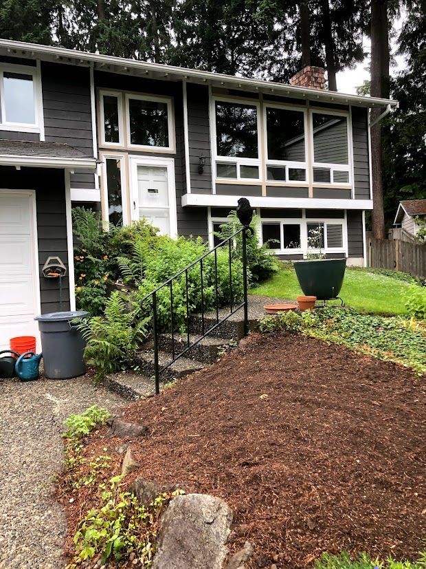 Dark gray house with large windows, front steps, and overgrown landscaping.