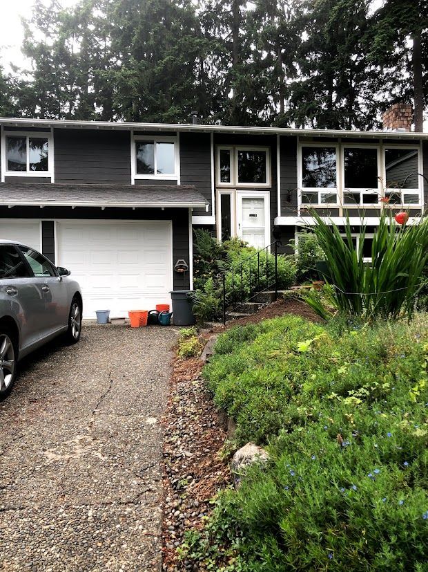 Gray house with white garage door and a car in the driveway, surrounded by greenery.