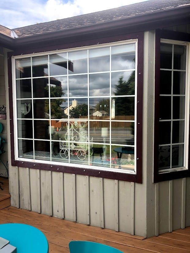 Exterior view of a house with large grid windows, brown trim, and a wooden deck with a blue table.