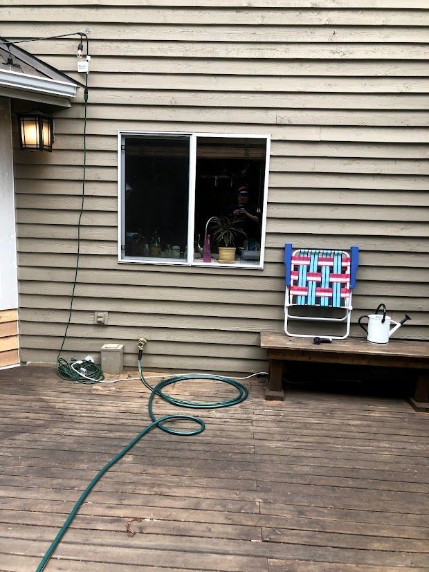 A wooden deck with a house and window; a hose, a folding chair, and a watering can.
