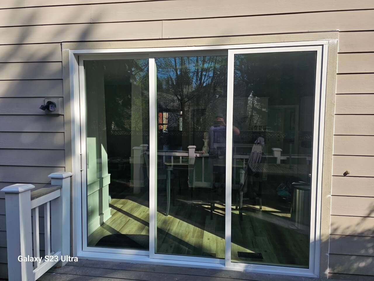 Sliding glass door with white frame, on a deck, reflecting a room with furniture and a person.