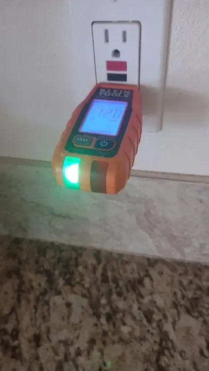 A voltage tester plugged into a white wall outlet displays a reading on its screen, with a green light illuminated.