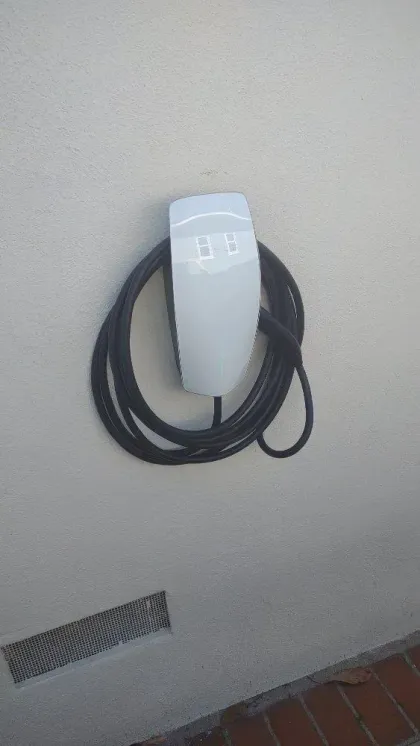 White electric vehicle charger mounted on a light-colored wall with a coiled black cable. Below is a vent.