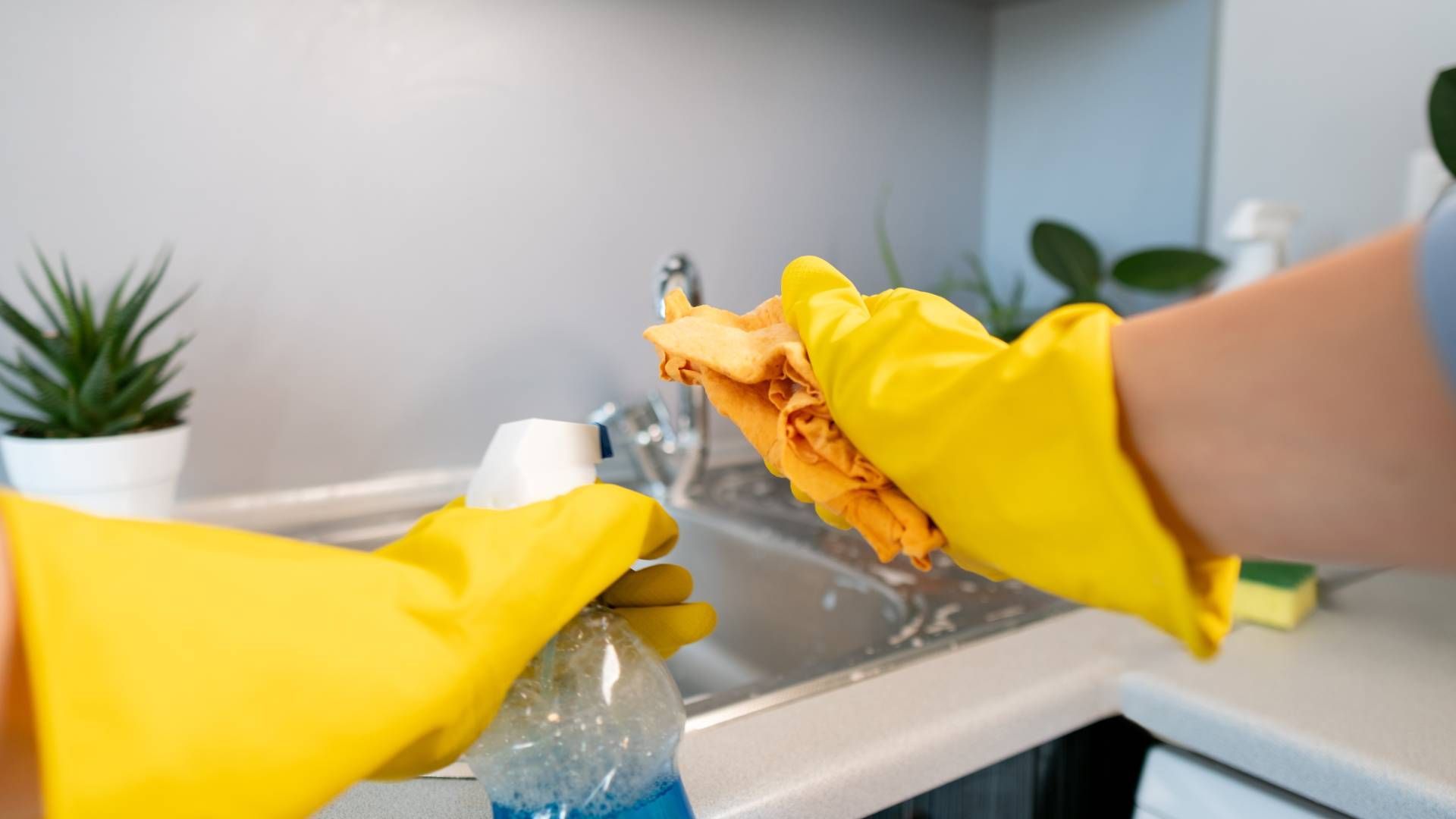 Person wearing yellow gloves cleaning a kitchen sink with a sponge and spray bottle.