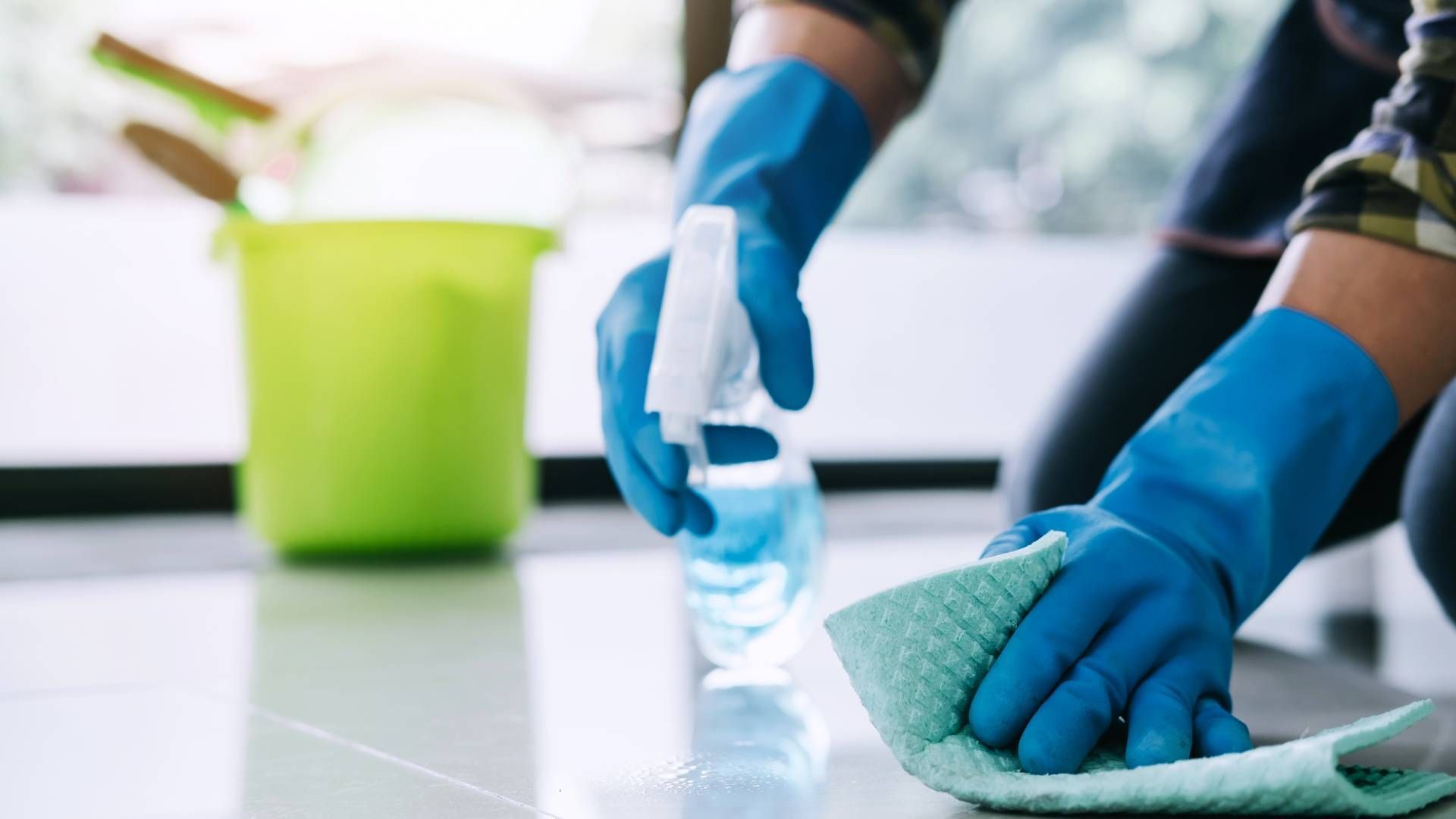 Person wearing blue gloves cleaning floor with spray bottle and cloth, bucket in background.
