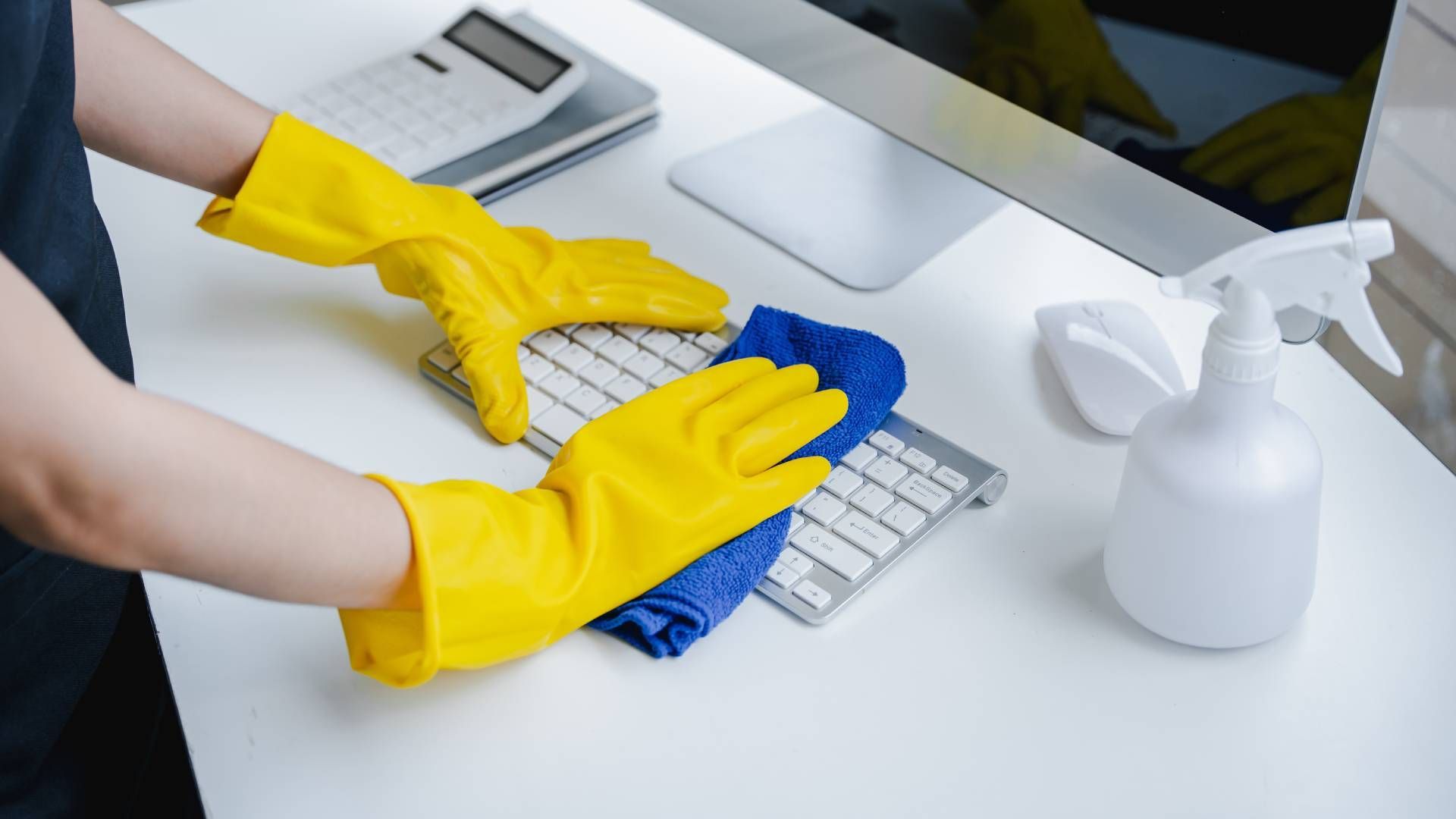 Person wearing yellow gloves cleaning a white desk with a blue cloth and spray bottle.