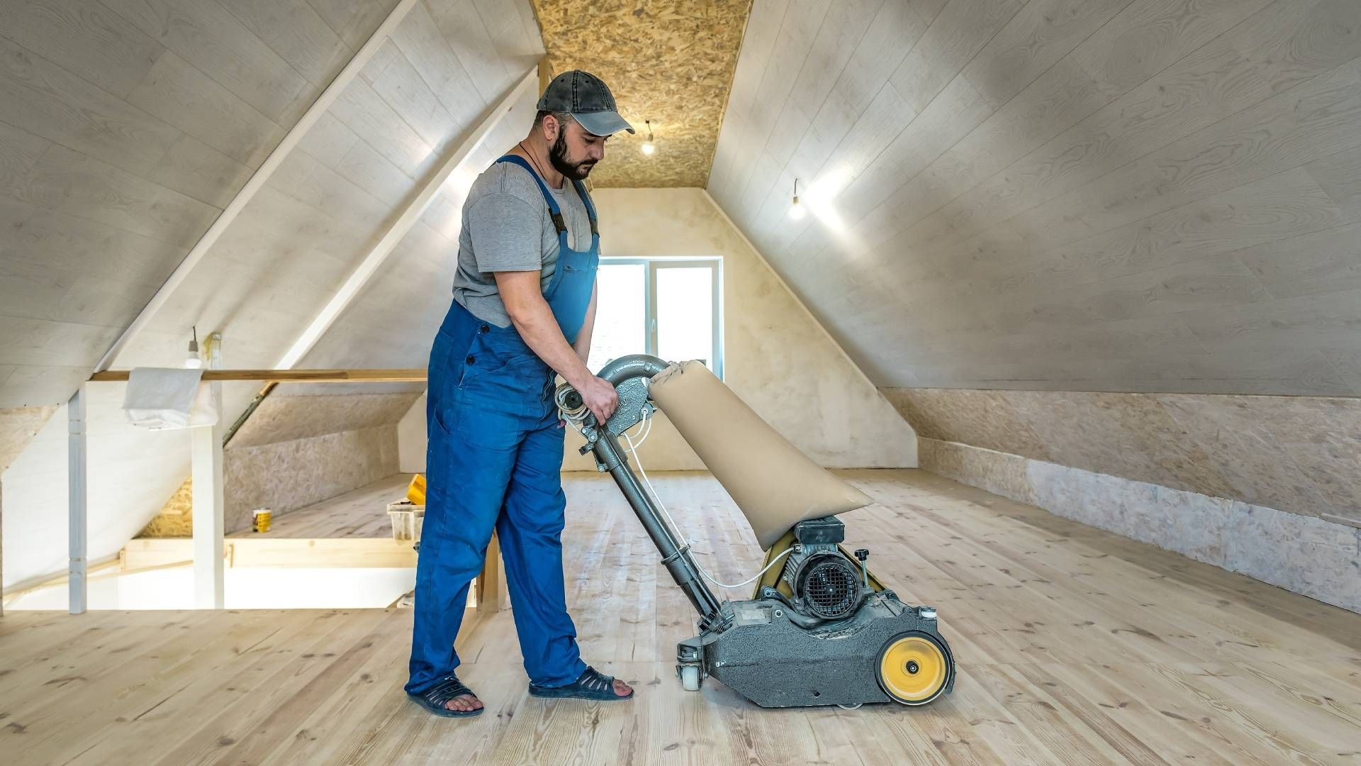 Man sanding wooden floor in an attic wearing blue overalls and a cap.