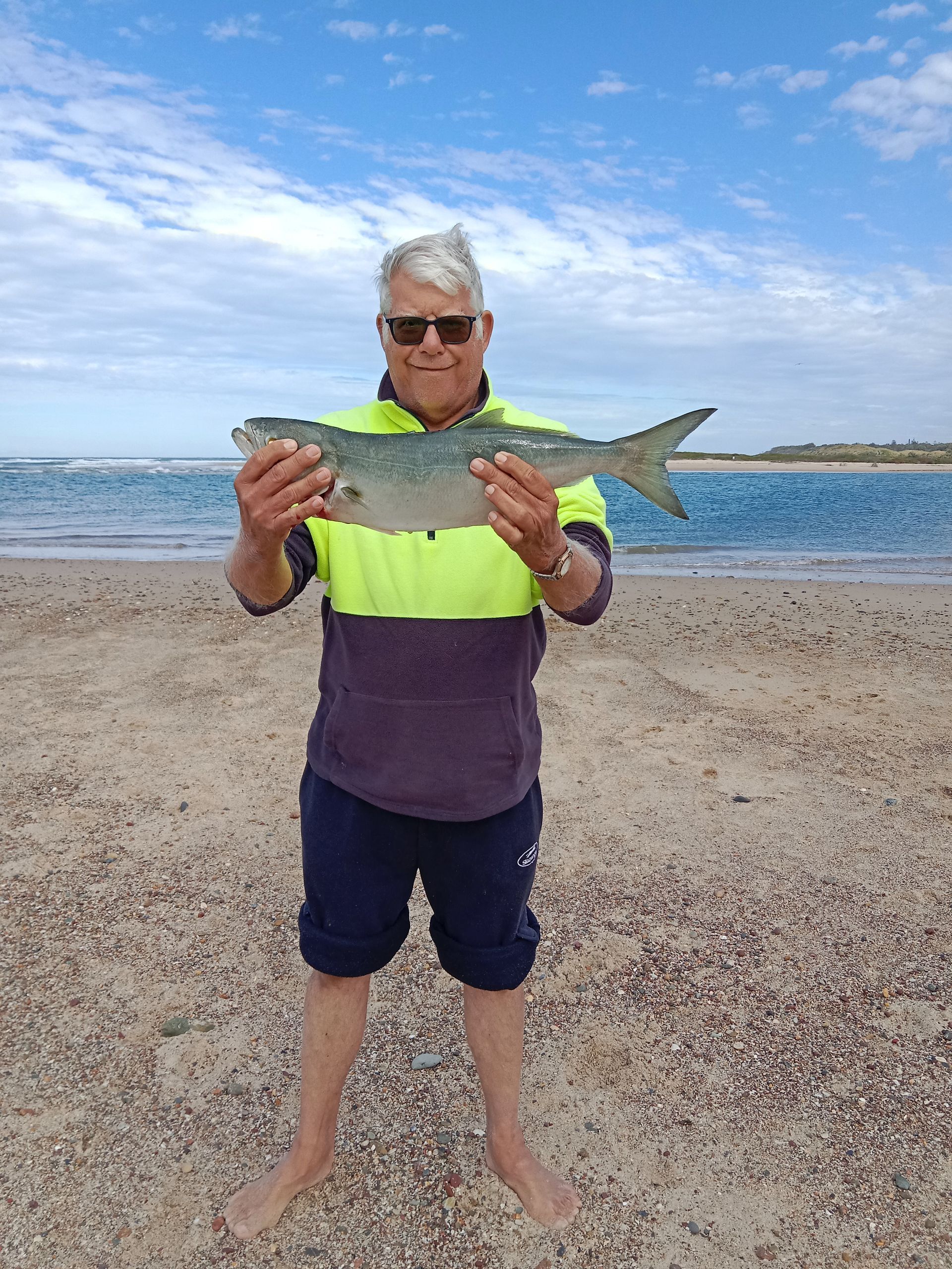 Man on beach holding a silver fish, blue water, and sky in the background. — VAL Disability Services in Taree, NSW