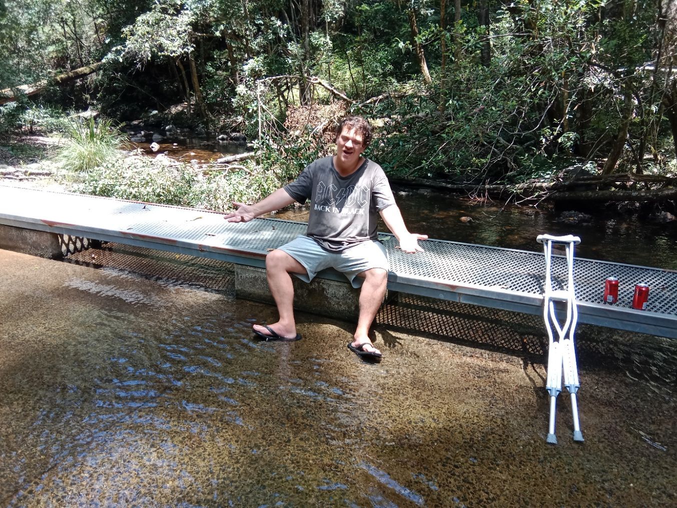 Man sitting with crutches on a bridge over water, arms outstretched, two cans on the right. — VAL Disability Services in Taree, NSW