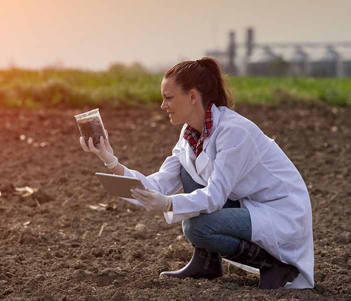 A woman in a lab coat is kneeling down in a field holding a tablet and a jar.