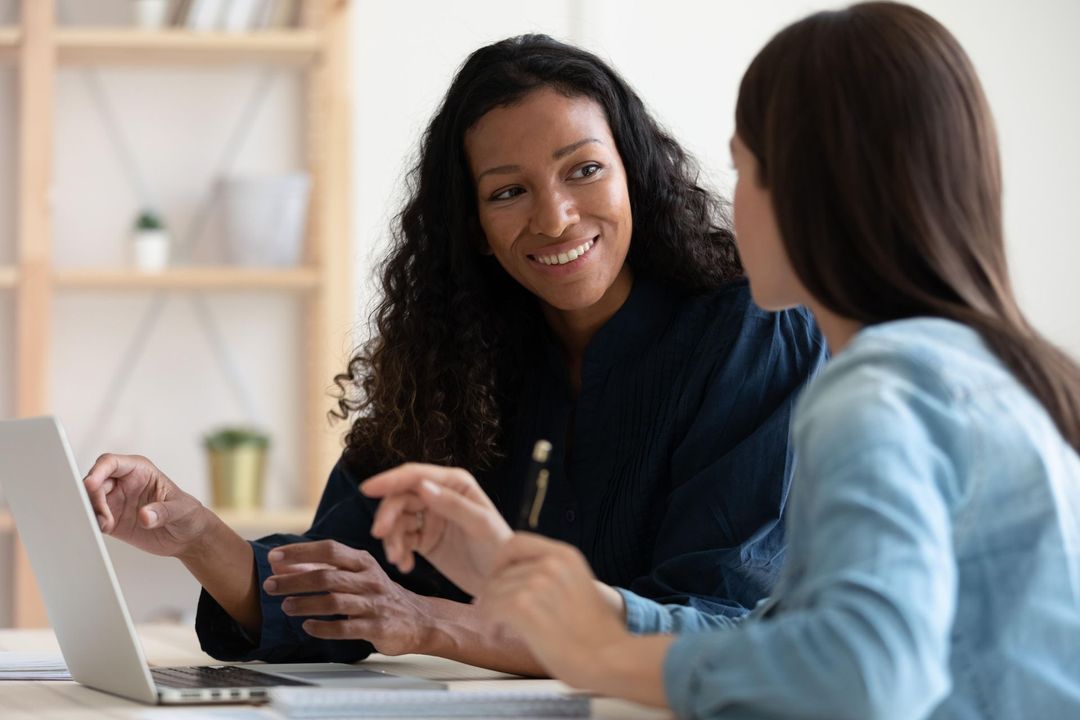 two women are sitting at a table looking at a laptop computer