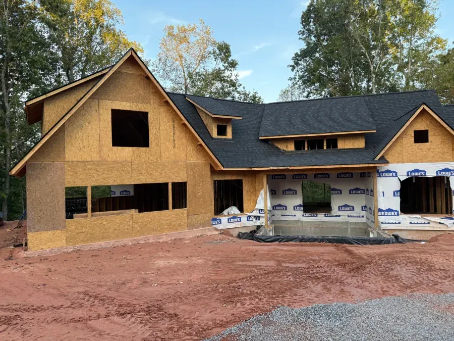 Partially built house with plywood siding, black roof, and red dirt yard surrounded by trees