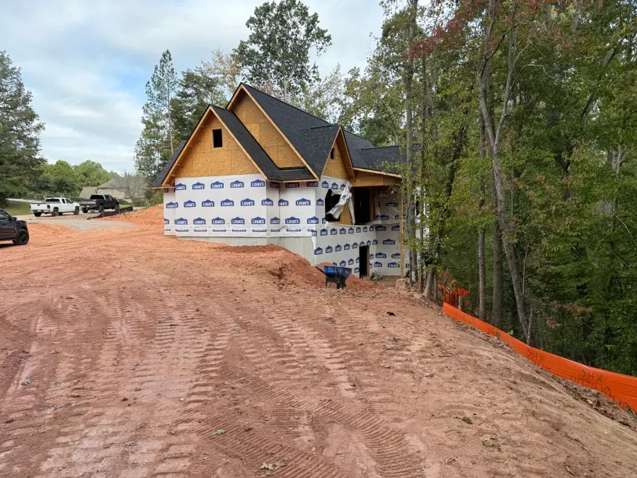 Partially built house with white wrap, red dirt driveway, and trees beside it