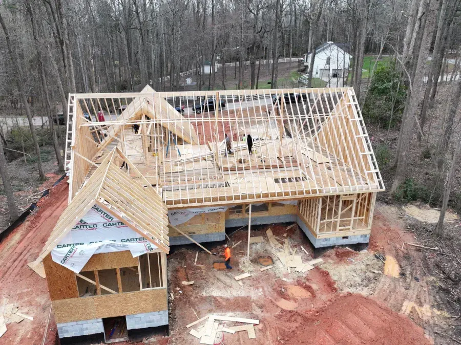 Partially framed house under construction with exposed roof trusses and workers inside, surrounded by trees