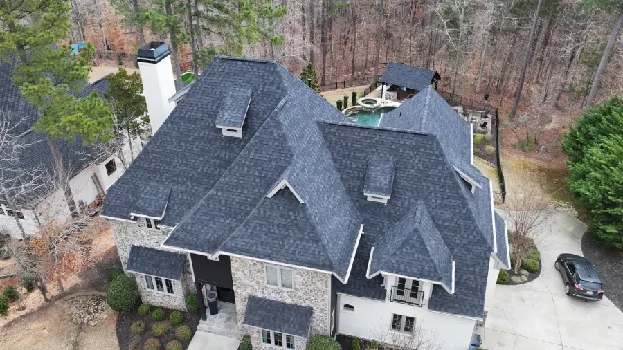 Aerial view of a large gray-roofed suburban house with a driveway and wooded backyard.