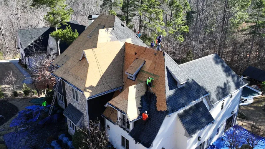 Aerial view of a house roof under repair, with workers and materials visible among surrounding trees.