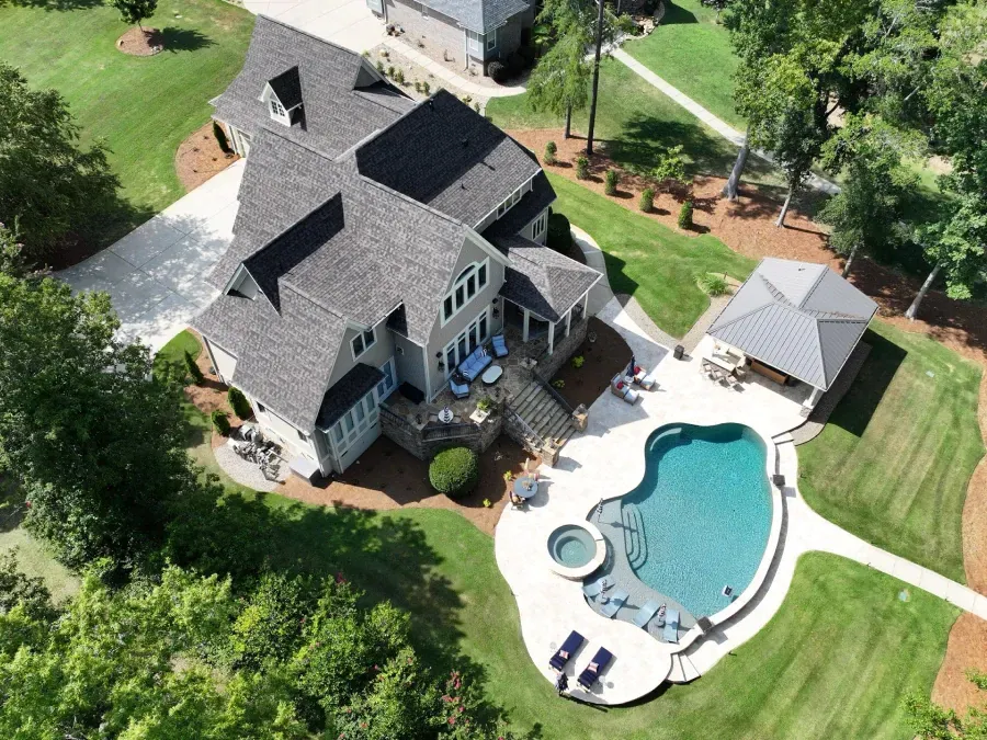 Aerial view of a large gray house with a backyard pool, patio, and detached garage surrounded by trees.