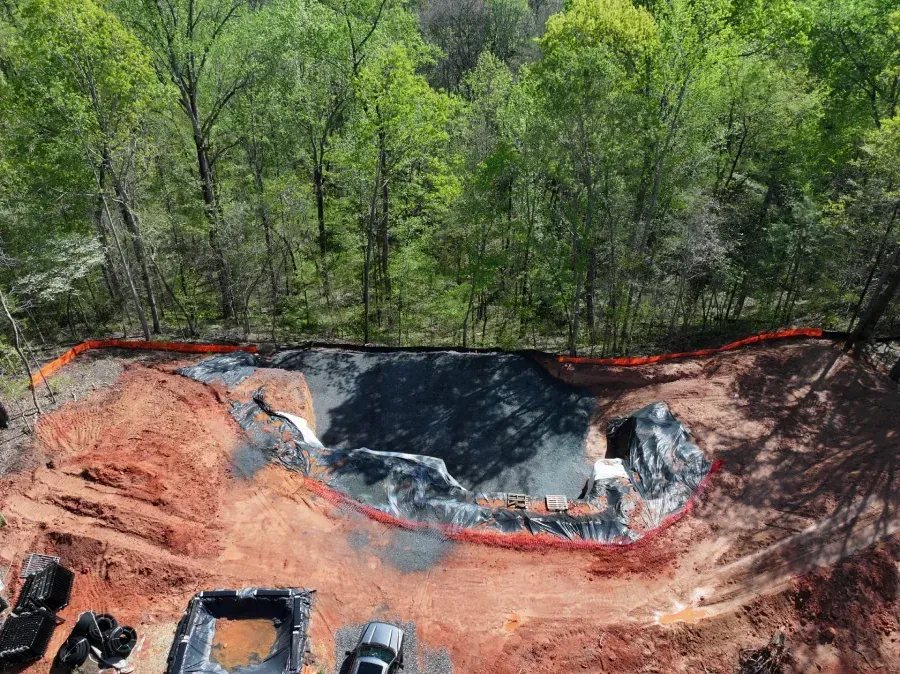 Large black-lined excavation pit in a wooded area, with orange soil and construction equipment at the bottom.