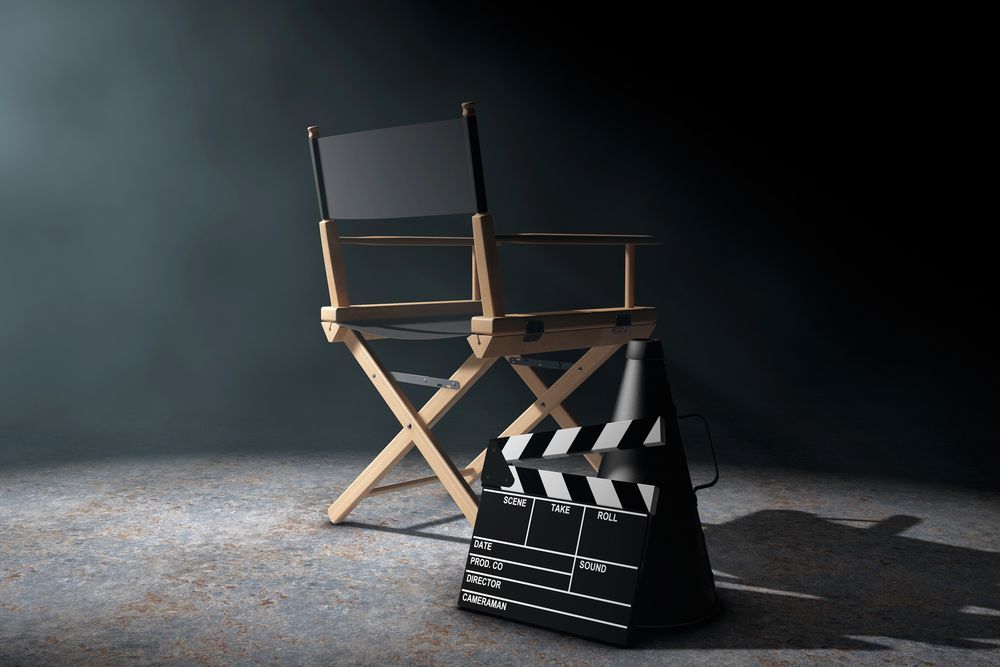 Una sedia da regista e un ciak cinematografico, sullo sfondo di un'ambientazione scura illuminata da faretti drammatici.