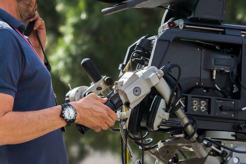 Un operatore di ripresa professionista regola una telecamera mentre parla al telefono, all'aperto, con uno sfondo sfocato.