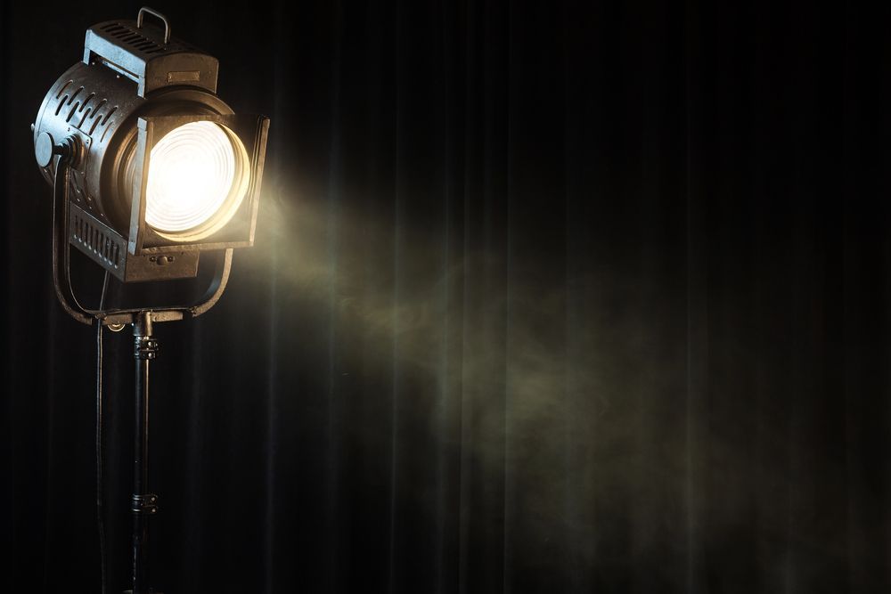 Un riflettore teatrale su un supporto inclinato verso destra proietta un fascio di luce intensa su una tenda nera.
